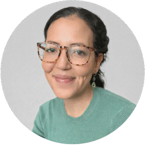 Woman with curly dark hair, tortoiseshell glasses, and a green top, smiling at the camera against a light gray background—an image that embodies the welcoming spirit of Brooklyn Therapy.