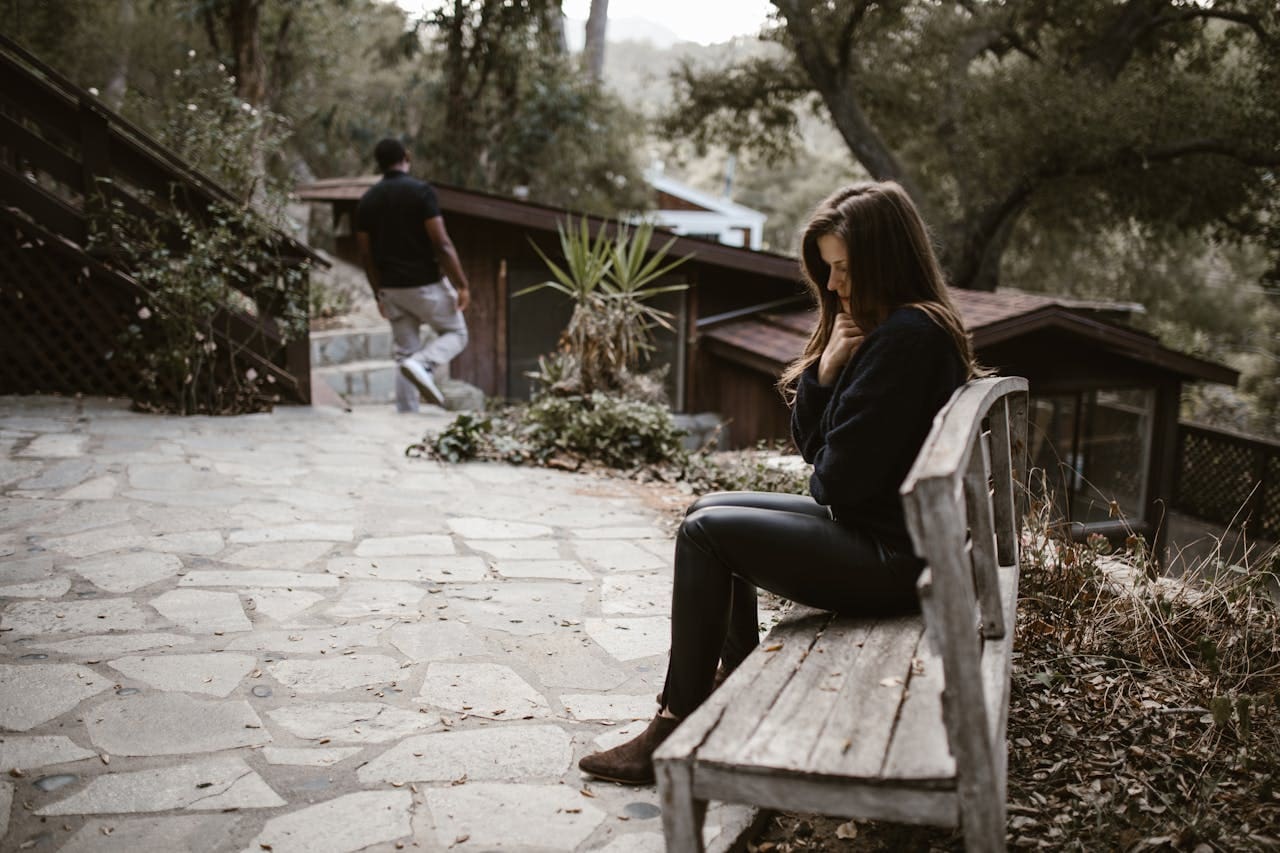 A woman sits on a wooden bench looking down, perhaps reflecting on her thoughts after a Brooklyn Therapy session, while a man walks away on a stone path in an outdoor setting with trees and rustic buildings.