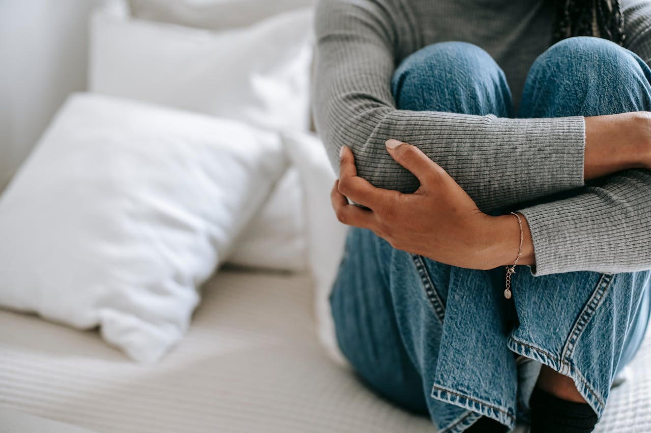 Person in a gray sweater and blue jeans sitting on a bed, hugging their knees—an image that captures the quiet moments often explored in therapy. White pillows are in the background, creating a calm setting reminiscent of Brooklyn Therapy sessions focused on comfort and self-reflection.