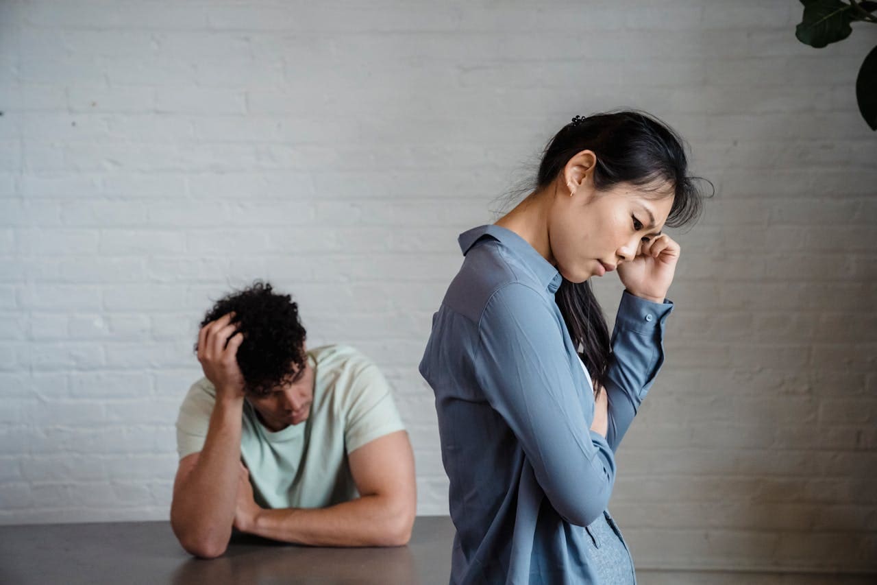 A woman stands with a hand on her forehead looking upset, while a man sits at a table behind her with his head in his hand, both appearing distressed—capturing a moment that underscores the importance of seeking support through therapy or connecting with a therapist NYC for guidance during difficult times.