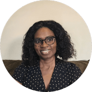 Woman with curly black hair and glasses, wearing a black polka dot shirt, sits on a couch against a plain light-colored wall, smiling at the camera—embodying the warm, approachable spirit often found in Brooklyn Therapy sessions with therapist NYC professionals.