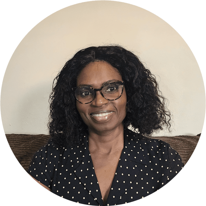 Woman with curly black hair and glasses, wearing a black polka dot shirt, sits on a couch against a plain light-colored wall, smiling at the camera—embodying the warm, approachable spirit often found in Brooklyn Therapy sessions with therapist NYC professionals.
