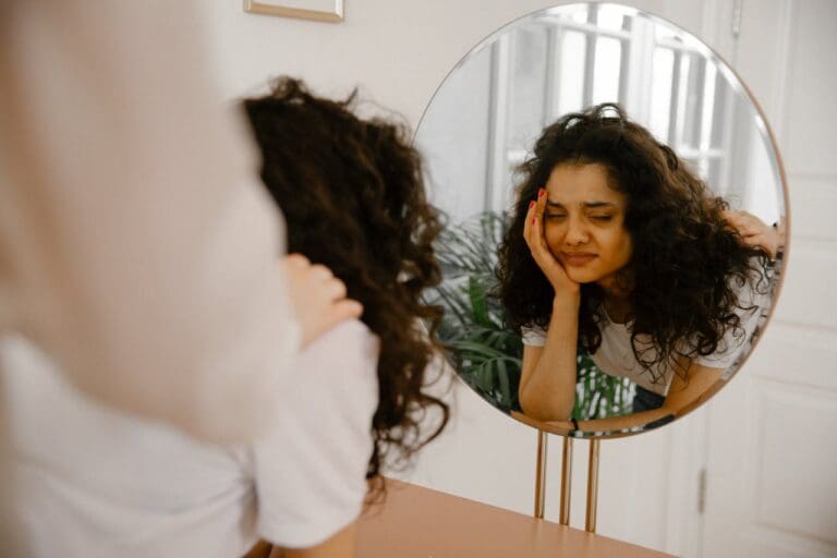 A person with curly hair looks at their reflection in a round mirror, resting their head on their hand and appearing tired or stressed—capturing a moment that might resonate with anyone considering therapy or seeking support from Brooklyn Therapy.