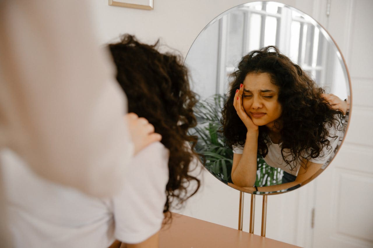 A person with curly hair looks at their reflection in a round mirror, resting their head on their hand and appearing tired or stressed—capturing a moment that might resonate with anyone considering therapy or seeking support from Brooklyn Therapy.