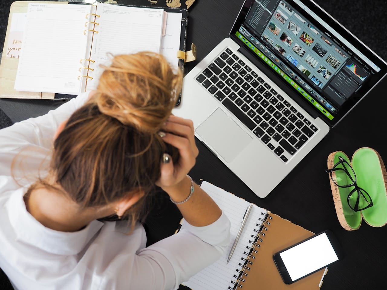 A person sits at a desk with a laptop, notebooks, planner, phone, and glasses, holding their head and looking stressed or overwhelmed while working—perhaps considering reaching out for therapy or connecting with a therapist NYC for support.