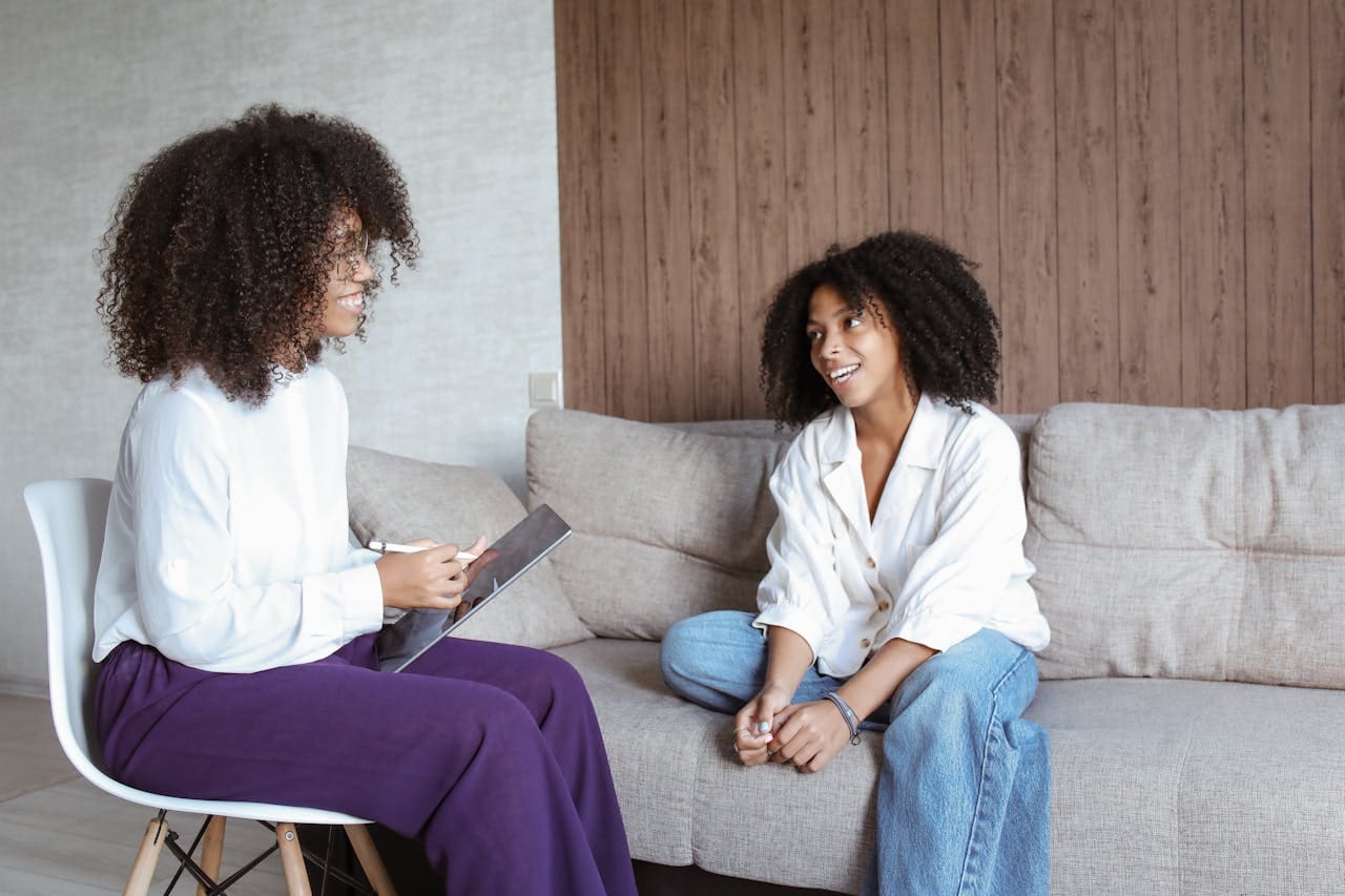 Two women sit indoors, one on a chair holding a tablet and a pen, the other on a couch, engaged in a conversation that suggests the supportive environment of Brooklyn Therapy.