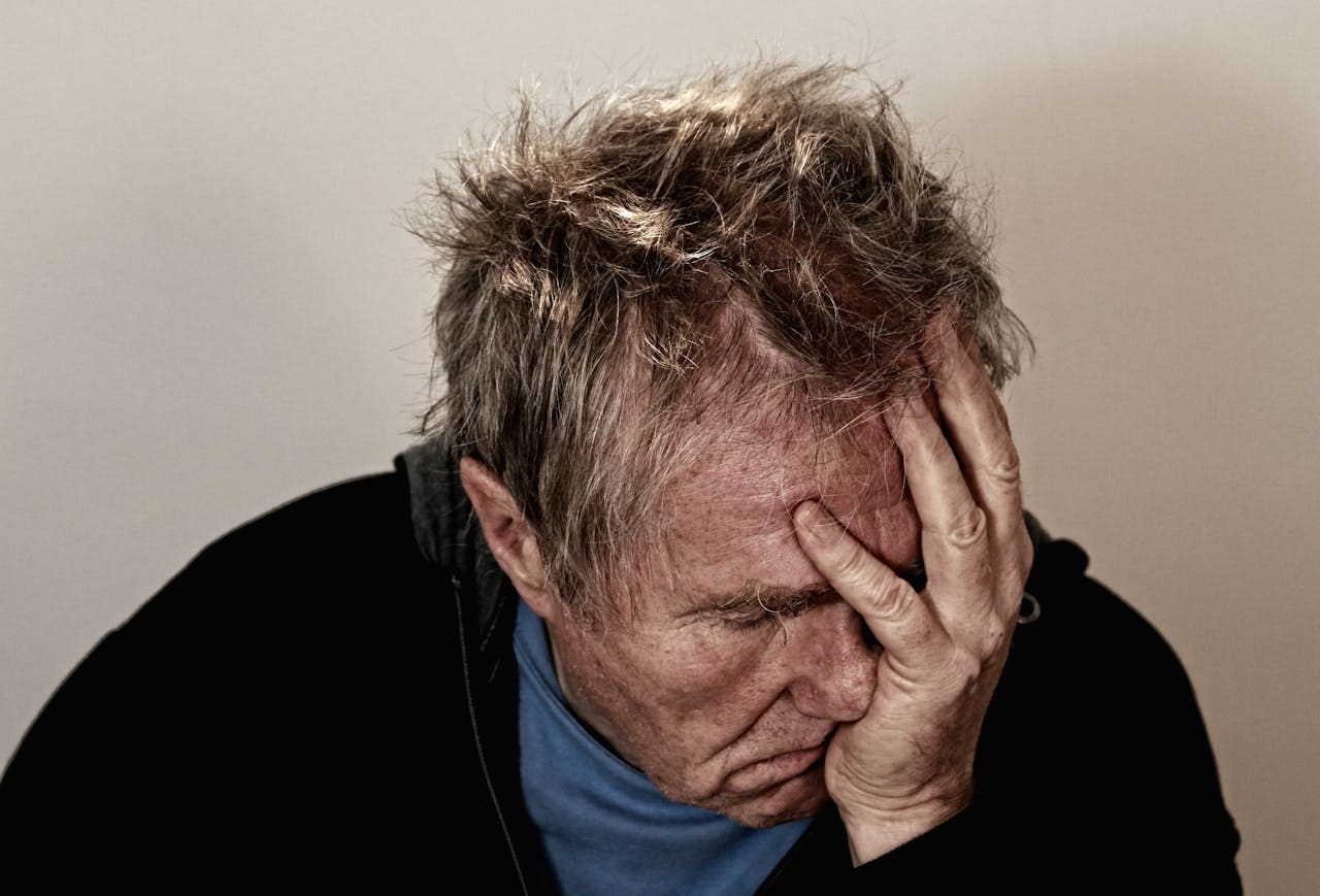 An older man with messy hair sits against a plain background, covering his face with one hand and appearing distressed or tired, as if searching for comfort through therapy or the support of a therapist in NYC.