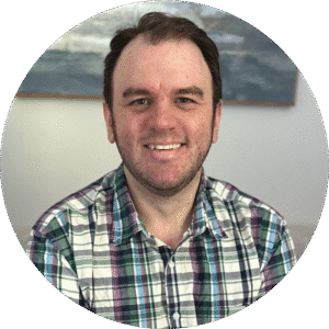 Man with short brown hair and a beard, wearing a plaid shirt, smiling at the camera with a blurred painting in the background—a warm image that reflects the approachable environment of Brooklyn Therapy, where your search for a therapist NYC can begin.