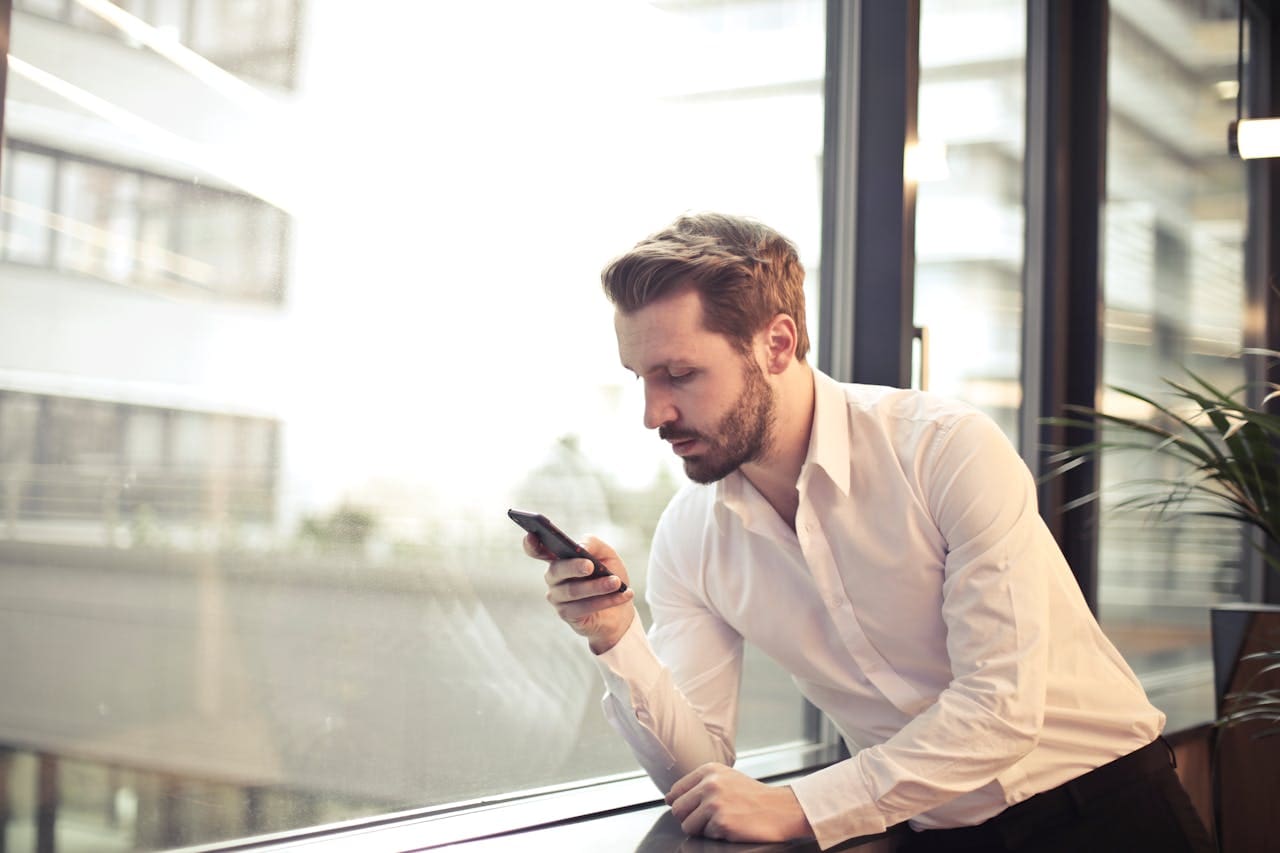 A man in a white dress shirt leans on a window ledge indoors, looking at his smartphone with a focused expression—perhaps searching for Brooklyn Therapy or connecting with a therapist NYC offers.