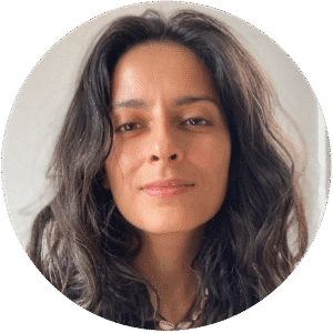 Woman with long wavy dark hair and a neutral expression, looking directly at the camera against a plain light background—reflecting the calm, approachable presence often sought in a therapist NYC or Brooklyn Therapy setting.