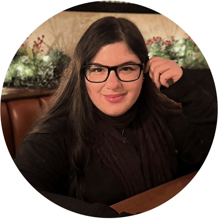 A person with long dark hair and glasses sits at a wooden table, smiling, with festive greenery and red berries in the background—creating a warm and welcoming space reminiscent of Brooklyn Therapy or a comforting therapy session in NYC.