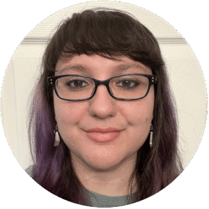 A person with long brown hair, glasses, and earrings is facing the camera and smiling slightly. The background is a plain white wall, evoking the calm and welcoming atmosphere often found in Brooklyn Therapy sessions or with a compassionate therapist NYC.