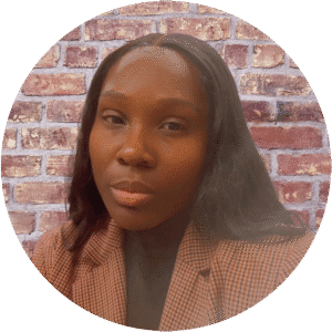 A person with long dark hair wearing a checked brown blazer, standing in front of a brick wall, exuding the calm confidence often associated with a Brooklyn Therapy professional.