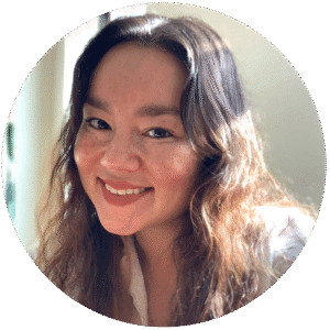 A woman with wavy brown hair smiles at the camera. She is wearing a white top and is in a softly lit indoor setting, creating a warm and welcoming atmosphere reminiscent of a Brooklyn therapy space.