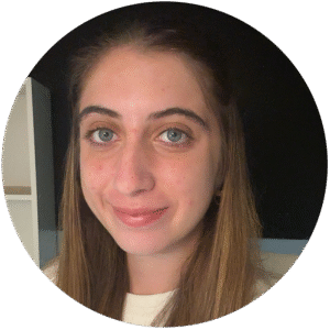 A woman with long brown hair and blue eyes is smiling slightly at the camera, standing indoors with a shelf and dark wall in the background, evoking a calm atmosphere often found in Brooklyn Therapy spaces.