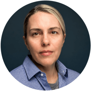 Person with light skin, short blonde hair, and a neutral expression, wearing a blue checkered shirt and gray undershirt, posed in front of a dark blue background—an approachable presence reflective of someone you might meet during Brooklyn Therapy sessions or while searching for a therapist in NYC.