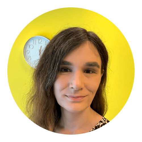 A person with long brown hair stands in front of a bright yellow wall with a round clock partially visible behind them, evoking the welcoming environment often found in Brooklyn Therapy or with a therapist NYC.