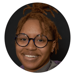 Person with short, reddish-brown locs, wearing round glasses and a nose ring, smiling against a black background—radiating the warmth and approachability you’d expect from a dedicated therapist NYC clients trust for compassionate Brooklyn therapy.