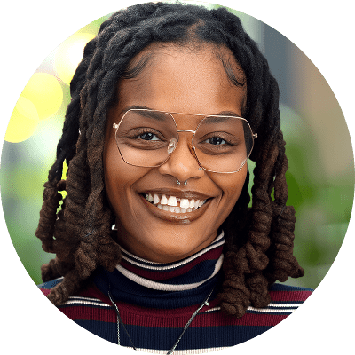 Smiling person with glasses and dreadlocks wearing a striped turtleneck, photographed in natural light with a blurred background—a welcoming face you might find at Brooklyn Therapy, where finding the right therapist NYC is made easier.