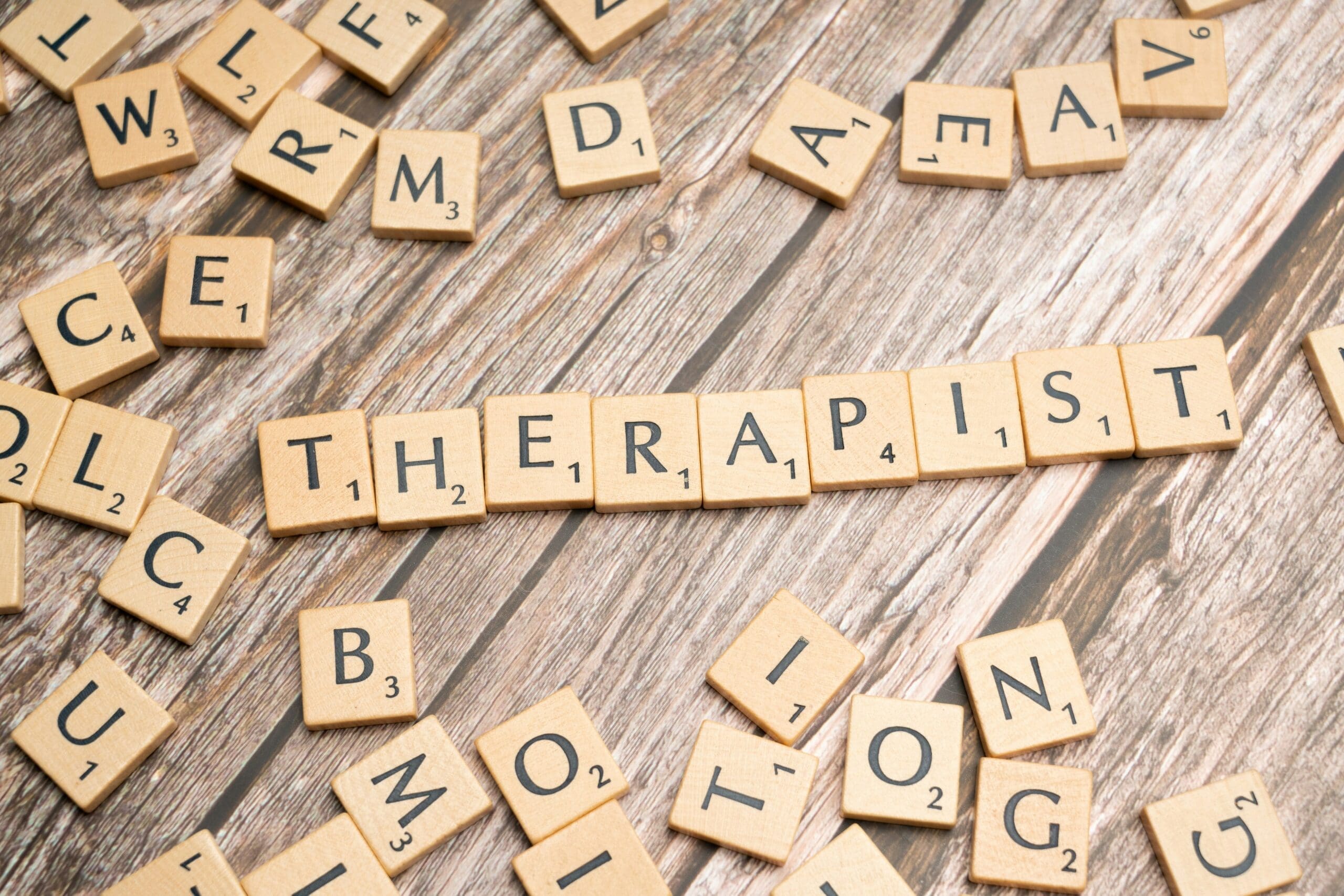 Scrabble tiles on a wooden surface spell “THERAPIST” in a row, surrounded by other scattered letter tiles, evoking the thoughtful process of therapy. This inviting scene captures the supportive environment fostered by Brooklyn Therapy and therapist NYC professionals.