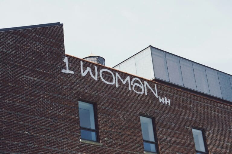 Graffiti reading “1 WOMAN” is painted in white on the upper brick wall of a building, reminiscent of the resilience often explored in Brooklyn Therapy sessions, with windows and a water tower visible in the background.
