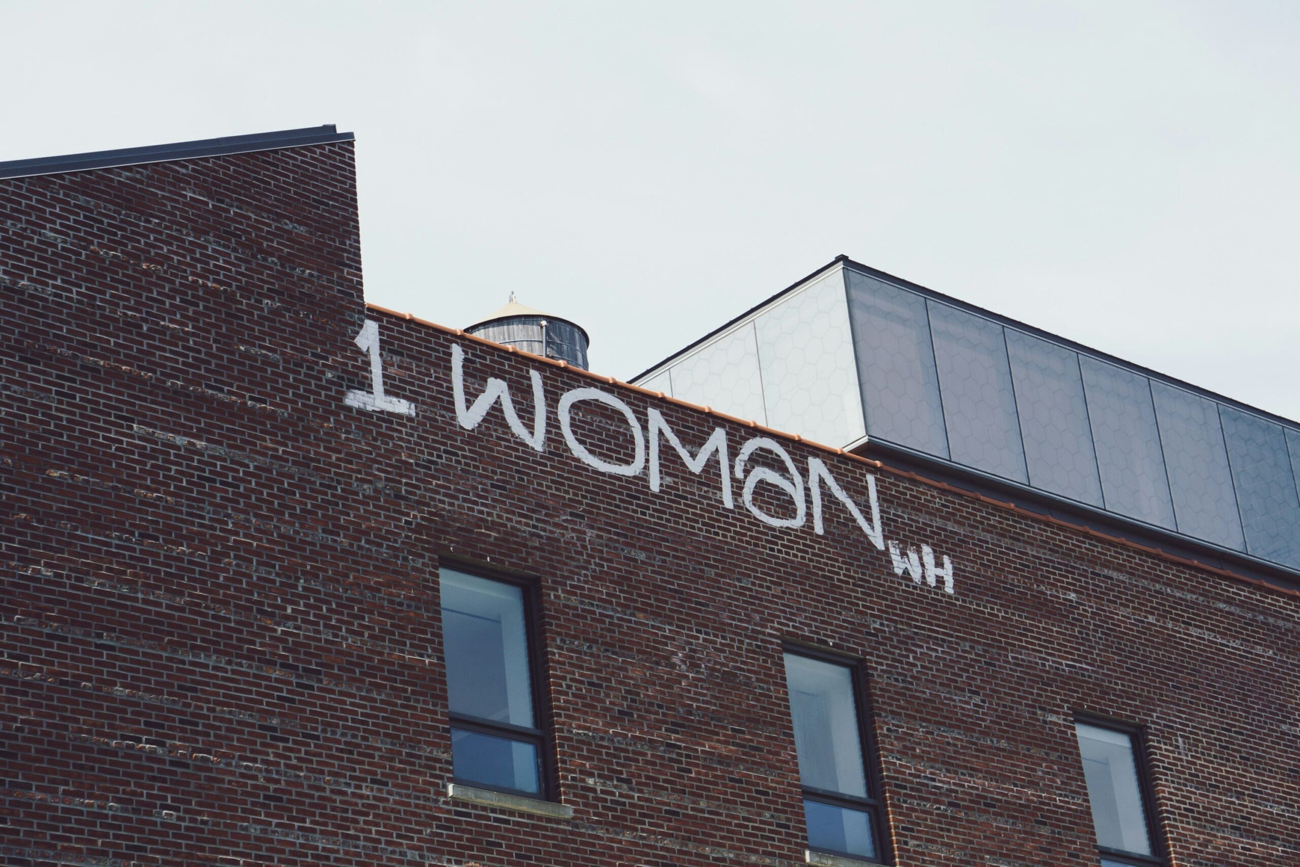 Graffiti reading “1 WOMAN” is painted in white on the upper brick wall of a building, reminiscent of the resilience often explored in Brooklyn Therapy sessions, with windows and a water tower visible in the background.
