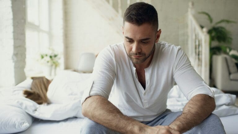 A man in a white shirt sits on the edge of a bed looking down, appearing thoughtful or upset, while a person lies in the background—capturing the kind of moments often explored in therapy sessions, such as those offered by Brooklyn Therapy.