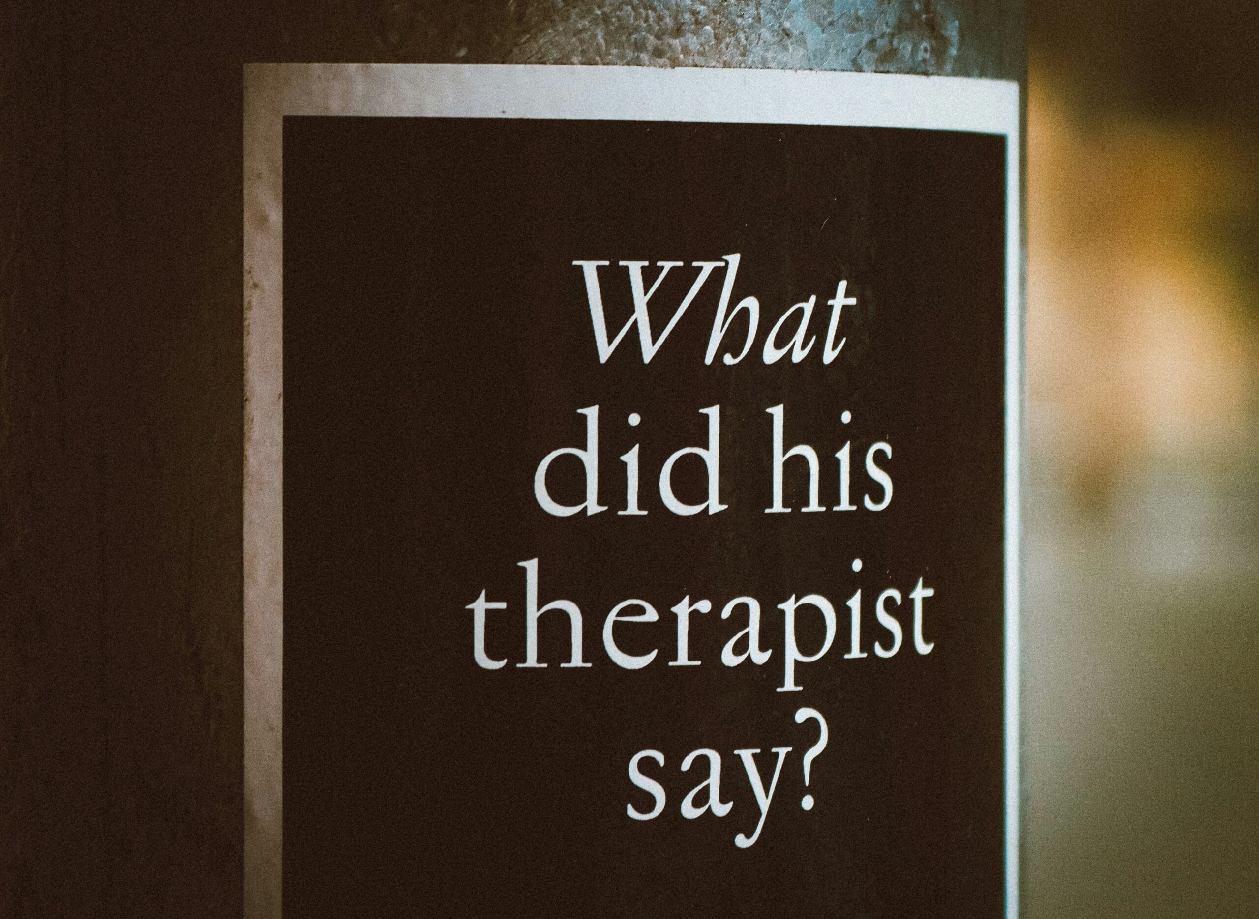 A black sign with white text reads, “What did his therapist say?” attached to a vertical surface—a playful nod to the culture of therapy in NYC and Brooklyn Therapy conversations.