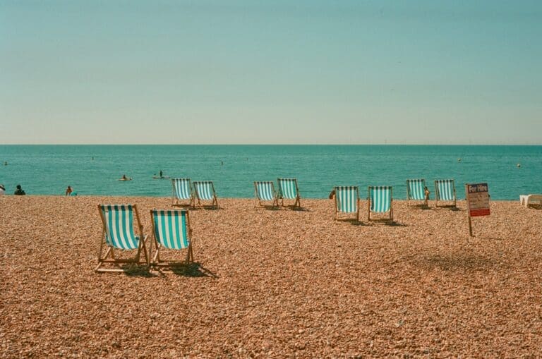 Empty striped deck chairs are arranged on a pebbly beach facing the calm sea under a clear sky, with a few people swimming in the distance and a sign visible on the right—an inviting scene reminiscent of the tranquil spaces created in Brooklyn Therapy, where calm and clarity meet.