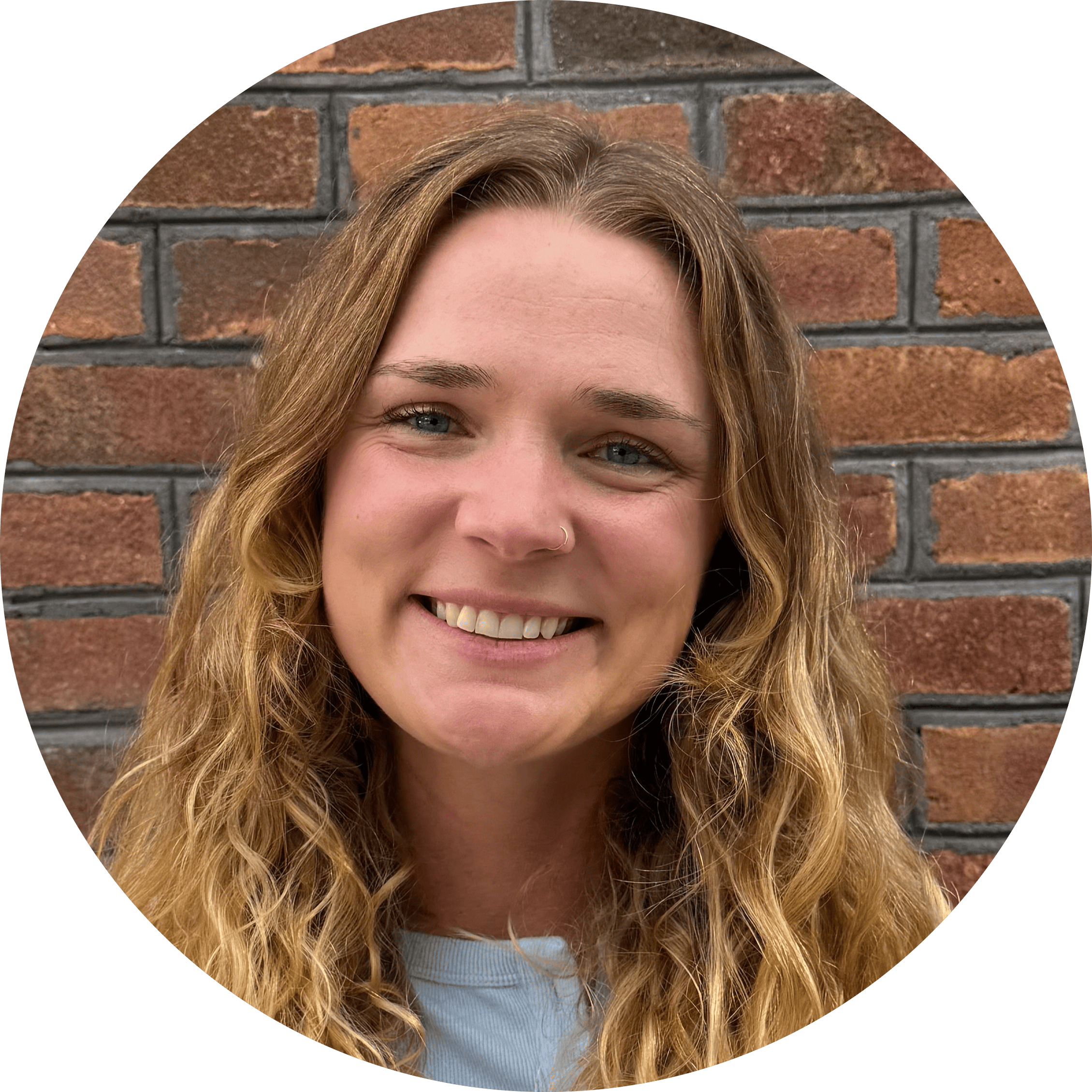 A woman with long wavy blonde hair smiles in front of a brick wall, wearing a light blue top—capturing the warm, approachable spirit you’ll find with Brooklyn Therapy.