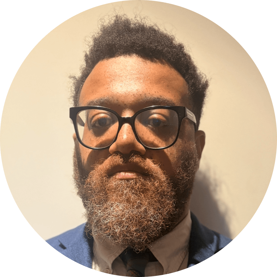 A person with short curly hair, a full beard, glasses, and a suit looks directly at the camera against a plain light background—capturing the approachable and professional presence you’d expect from a Brooklyn Therapy specialist or therapist NYC clients trust.