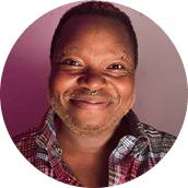 A person with short hair smiles at the camera, wearing a plaid shirt, against a pink and white background—a warm and welcoming image that captures the approachable spirit often found in Brooklyn Therapy.