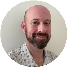 A bearded man with a bald head wearing a light-colored, patterned shirt smiles at the camera in front of a plain white background.