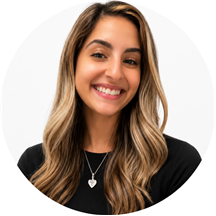 Smiling woman with long, wavy blonde hair wearing a black top and a heart-shaped pendant necklace, photographed against a plain white background.