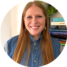 A person with long red hair and a denim shirt smiles in front of a bookshelf filled with various books.