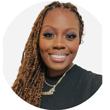 Woman with long, braided hair smiling at the camera, wearing a black shirt and gold necklaces, against a plain white background.