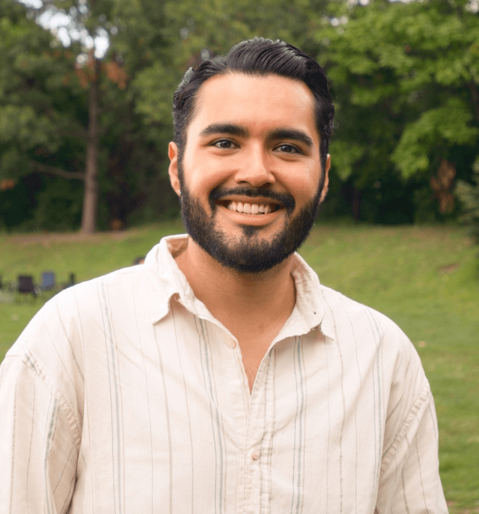 A man with a beard and mustache smiles outdoors, wearing a light-colored, button-up shirt. Green trees and grass fill the background.