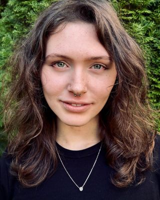 A person with wavy brown hair, a nose ring, and a necklace is standing outdoors in front of green foliage, looking directly at the camera.