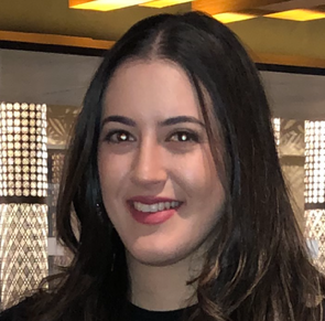 A woman with long dark hair and a black top smiles at the camera indoors, with modern lighting and a patterned wall in the background.